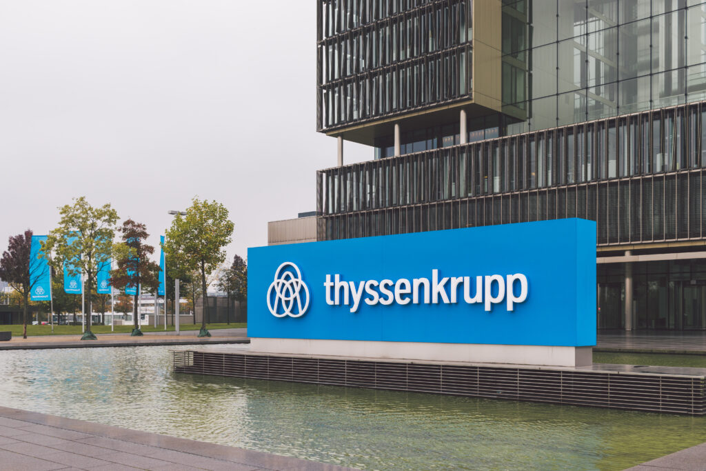 Thyssenkrupp Transformation Strategie: Thyssenkrupp-Logo vor Industriegelände mit Werksanlagen und Windkraftanlagen.